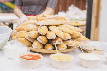 Ingredients for sandwiches, fresh hot baguettes, diffrent sauces on table, selective focus. Seller hands in background. Real scene in market.