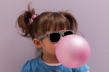 Little girl with sun glasses blowing up pink chewing gum against a light purple background