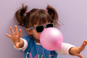 Little girl with sun glasses blowing up pink chewing gum against a light purple background