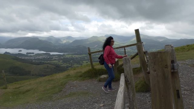 Cheerful long hair woman hiker coming to the fence gate on the hill in Lake District in England. Keswick town and Derwentwater in the valley in background.
