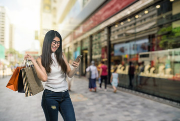 Fototapeta premium Happy young asian woman shopping in happy feeling and holding the product paper bag over Abstract blurred photo of shopping store in outdoor store mall with people walking streer, shopping concept