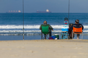 Two men sitting by the ocean fishing with a ship in the background