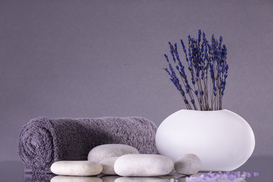 Spa. Lavender flowers stand in a white vase on a gray background. Pebbles and a towel rolled into a roll.