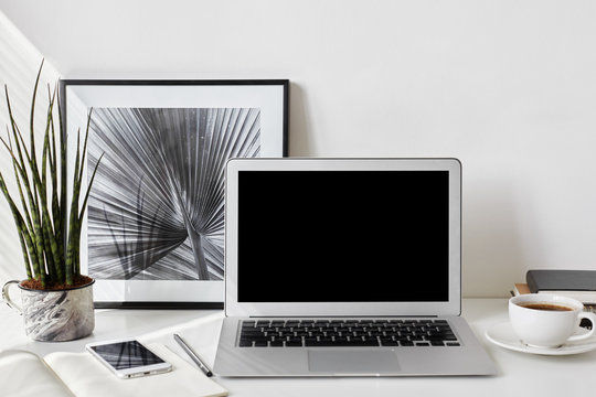 Set of modern electronic devices on white office desk. Freelancer's desktop with blank screen laptop, stationery items, framed picture, mobile phone, decorative plant and cup of coffee. Mock up