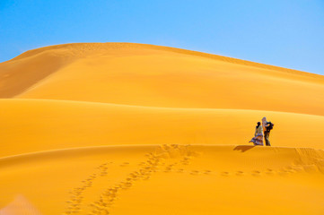 couple walks on high sandy dunes in bright sunny day, leave behind marks on sand, wind develops the girl's dress