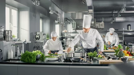Fotobehang Koken Beroemde chef-kok werkt met zijn hulp in een grote restaurantkeuken. De keuken staat vol met voedsel, groenten en kookgerei.