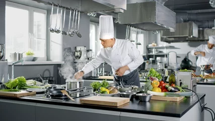 Selbstklebende Fototapeten Der berühmte Koch arbeitet mit seinen Lehrlingen in einer großen Restaurantküche. Die Küche ist voller Lebensmittel, Gemüse und kochendem Geschirr. © Gorodenkoff