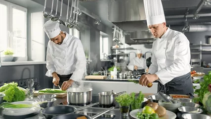 Türaufkleber Zwei berühmte Köche arbeiten als Team in einer großen Restaurantküche. Gemüse und Zutaten sind überall, Küche sieht modern aus mit viel Edelstahl. © Gorodenkoff