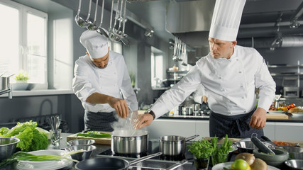 Zwei berühmte Köche arbeiten als Team in einer großen Restaurantküche. Gemüse und Zutaten sind überall, die Küche sieht modern aus mit viel Edelstahl.