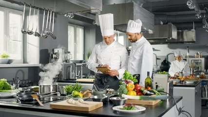 Photo sur Plexiglas Cuisinier Deux chefs célèbres discutent de leur blog vidéo tout en utilisant une tablette. Ils travaillent sur une grande cuisine professionnelle en acier inoxydable de restaurant.