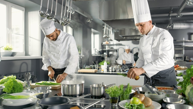 Two Famous Chefs Work as a Team in a Big Restaurant Kitchen. Vegetables and Ingredients are Everywhere, Kitchen Looks Modern with Lots of Stainless Steel.