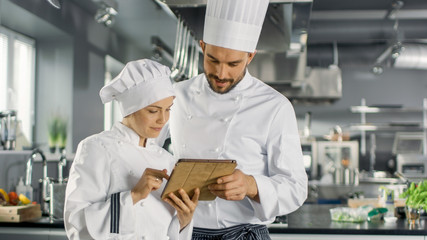 Male and Female Famous Chefs Discuss Their Video Blog while Using Tablet Computer. They Work on a Big Restaurant Stainless Steel Professional Kitchen.