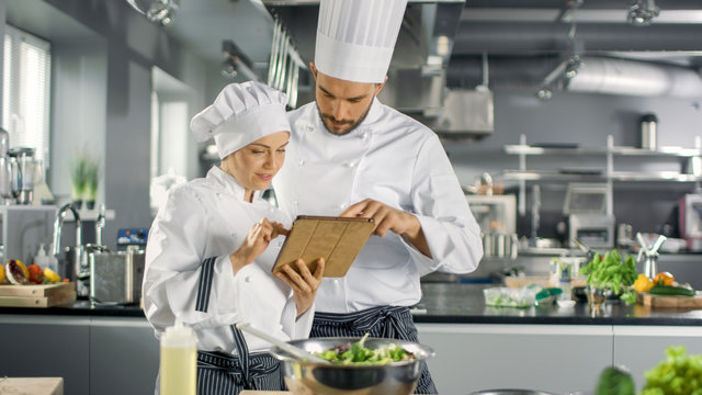 Male and Female Famous Chefs Discuss Their Video Blog while Using Tablet Computer. They Work on a Big Restaurant Stainless Steel Professional Kitchen.