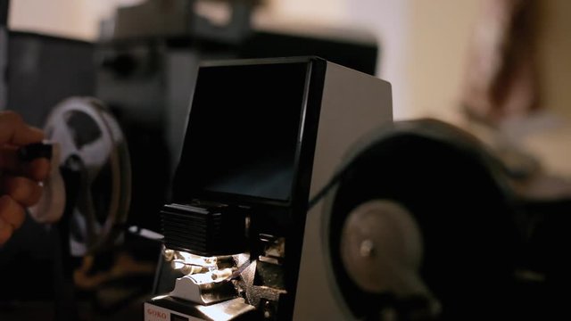 An editor moving the handles of a vintage film editing machine (a moviola, a device that allows viewing a reel on a tiny screen at a reduced speed), at a constant speed.
