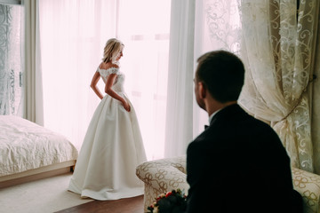 Young stylish groom sits in the chair and looking for his bride. Beautiful blonde near a window on the background of the stylish room.