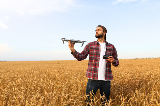 Bearded hipster man shows small compact drone and holds remote controller in his hand. Farmer agronomist looks at quadcopter before the launch in a wheat field
