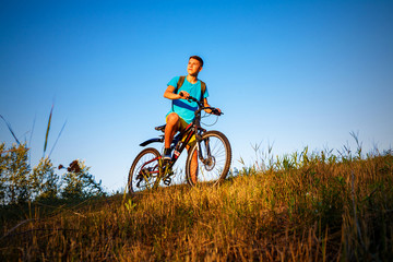 The boy on a bicycle is looking the sunset. The boy is standing on the hillside, behind him a blue sky without clouds.