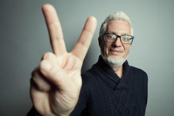 Nutty professor, old hipster concept. Portrait of 60-year-old man standing over gray background and showing V sign. Close up. Studio shot