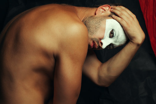Arty portrait of circus performer holding his head with make-up like venetian mask (volto bianco), posing over black, red curtains. Muscular body and perfect tan. Studio shot