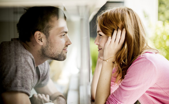 A couple smiling at each other through a pane of glass of a window.