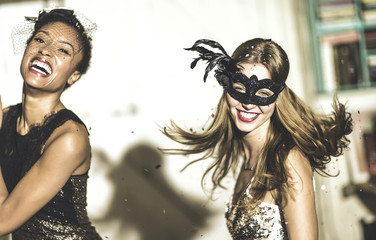 Two young women at a party in sequined dresses drinking and laughing, one wearing a face mask. 