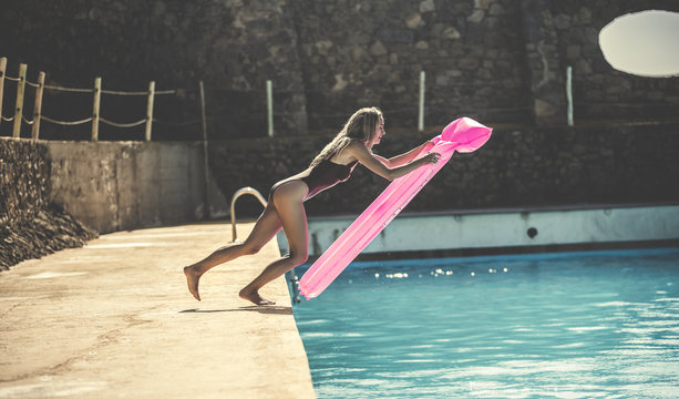 A young woman wearing a swimsuit jumping into a swimming pool holding a pool raft.