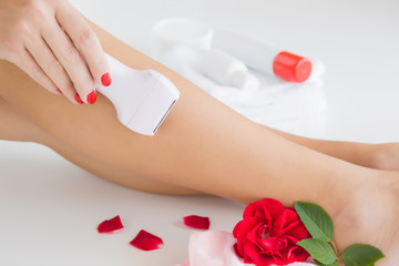Obraz na płótnie Canvas Woman epilates her leg with an electric epilator device. Smooth skin. Body care with soft cream. Summer atmosphere with fresh and fragrant red rose.
