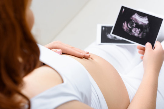 cropped view of pregnant woman touching her belly and holding ultrasound scans at home