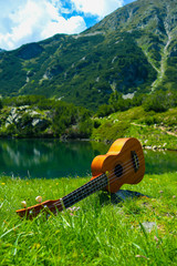 Romantic view of the Ukulele guitar at the mountain nature green meadow. Photo depicts musical instrument Ukulele small guitar at the amazing pure mountain lake Shore and the blue sky background. - 166230029