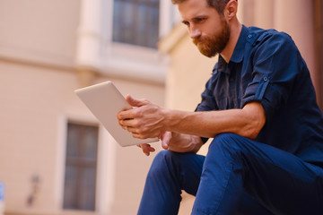 Bearded male sits on a step and using a tablet.