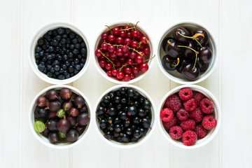  Bowls with fresh ripe gooseberry, red currant, black currant, raspberry, blueberry and cherry on white wooden table