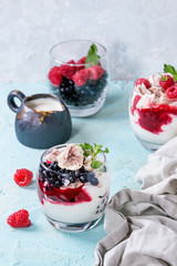 Traditional summer dessert Eton Mess. Broken meringue with whipped cream, berry jam, fresh blueberries and raspberries in two glasses, decorated with mint leaves over light blue concrete background.