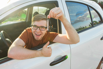 Naklejka premium Young driver showing car keys and thumbs up happy. Man holding car key for new automobile. Rental cars or drivers licence concept with male driving in beautiful nature on road trip.