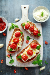 Tomato Ricotta Bruschetta with sun dried tomatoes paste, olive oil brown bread and basil in a white wooden board.