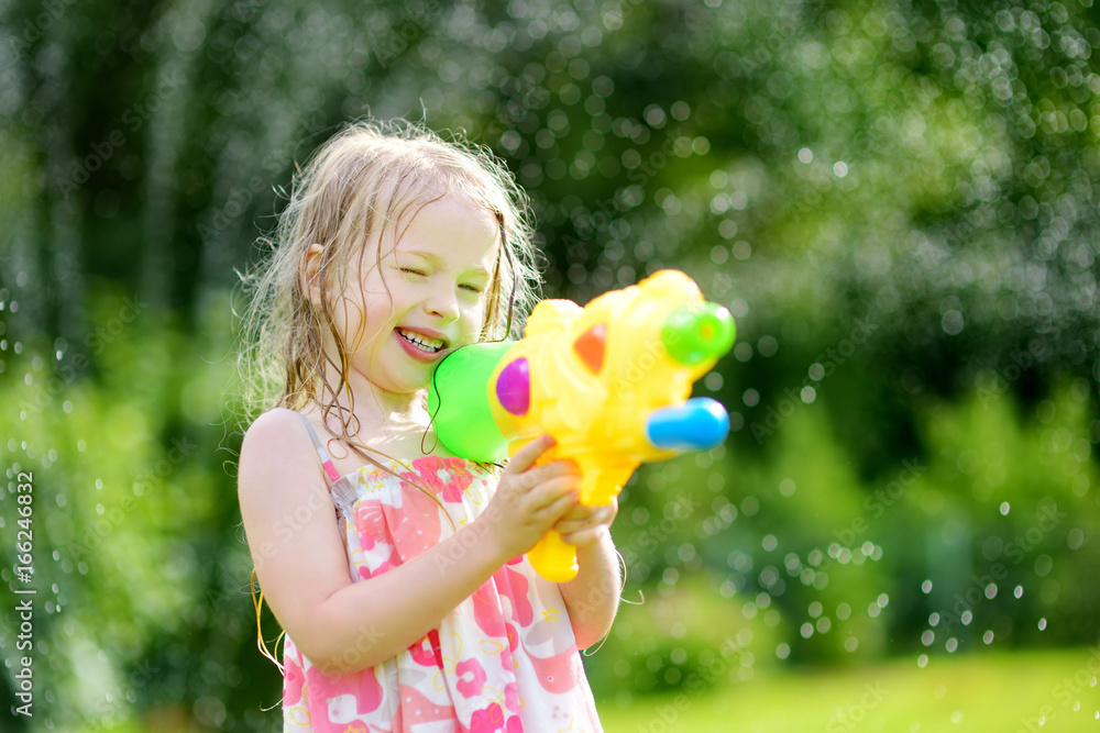 Sticker Adorable little girl playing with water gun on hot summer day. Cute child having fun with water outdoors.