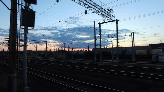 The train is approaching the railway station in the background of a sunset by the side of a freight train standing on the neighboring tracks.