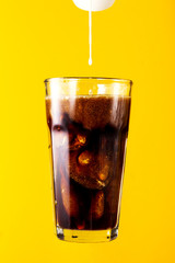Tasty fresh iced cold black coffee with ice and straw in glass pouring cream on yellow vivid background. Closeup.