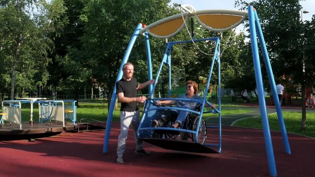 A sun with his mother in the swing. A lady in a wheelchair with her sun near her is sitting in the swing contracted specially for disabled persons while her sun is moving the swing to and fro