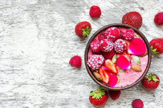 bowl of homemade pink smoothie with fresh raspberries, strawberries, nuts, seeds and roses flowers