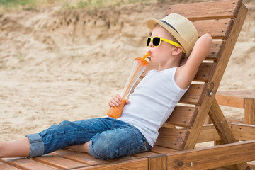 The boy in the straw hat and sunglasses  lying on the wooden sun lounger on the beach and drink fresh juice.Summer vacation.