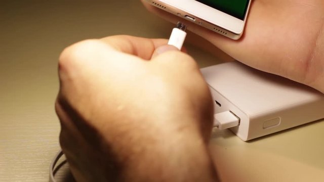 Hands of a male connect the USB cable to a white Power Bank (powerbank, batery) and phone
