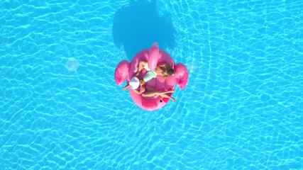 AERIAL TOP DOWN Young couple toasting and drinking cocktails while lying on fun inflatable flamingo floatie. Sexy couple in swimsuit sipping drinks and relaxing on pink flamingo floating on pool water - Powered by Adobe