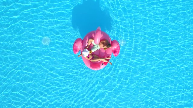 AERIAL TOP DOWN Young couple toasting and drinking cocktails while lying on fun inflatable flamingo floatie. Sexy couple in swimsuit sipping drinks and relaxing on pink flamingo floating on pool water
