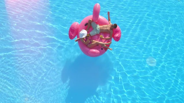 AERIAL LENS FLARE: Relaxed couple lying on fun inflatable flamingo floatie, drinking cocktails and relaxing. Sexy couple in swimsuit sipping drinks and enjoying on pink flamingo floating in sunny pool