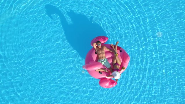 AERIAL: Young couple drinking cocktails and relaxing while lying on fun inflatable flamingo floatie. Sexy couple in swimsuit sipping drinks and enjoying on pink flamingo floating on sunny pool water