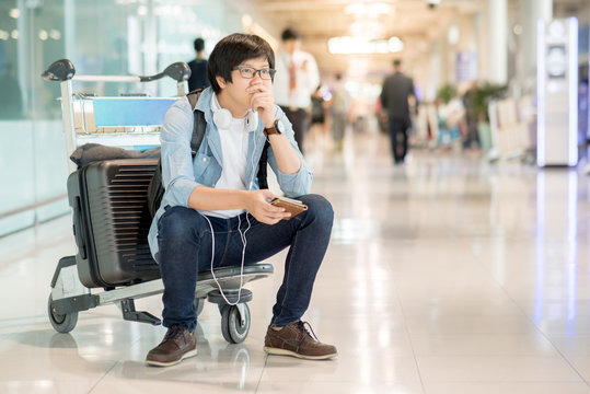 Young Asian Man Feeling Exhausted Sitting On Airport Trolley With His Suitcase Luggage In The International Airport Terminal, Flight Problem And Travel Insurance Concepts