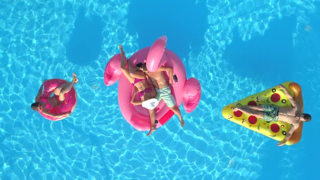 AERIAL TOP DOWN Smiling girls and guys in swimsuits laying on fun inflatable pizza, doughnut and flamingo floating on water. Happy friends enjoying on pink flamingo, doughnut and pizza floats in pool