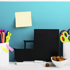 School background: a white brick wall with accessories, pencils, tetrads, scissors, an apple, pecans, a board for a substitute placed in front of her on the table. Selective focus.