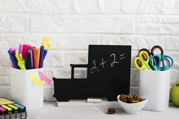 School background: a white brick wall with accessories, pencils, tetrads, scissors, an apple, pecans, a board for a substitute placed in front of her on the table. Selective focus.