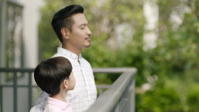 Asian father & son standing on balcony, smiling, looking at each other and taking deep breath
